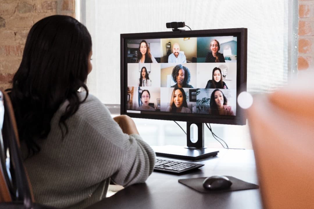 Woman in grey sweater attends a video conference with nine participants on a computer monitor.