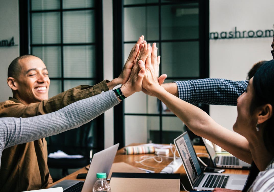 Diverse colleagues smiling and giving a high-five over their laptops in a modern office.